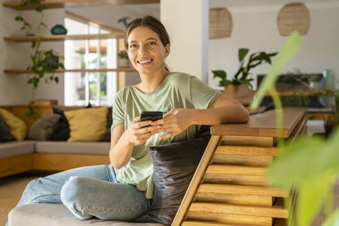 Lächelnde junge Frau mit Smartphone sitzt zu Hause auf dem Sofa - SBOF03230
