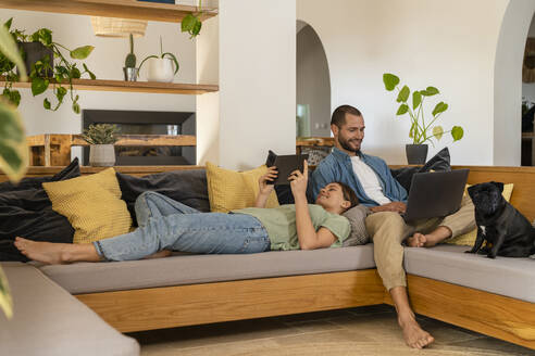 Glückliches junges Paar, das ein digitales Tablet und einen Laptop benutzt, während es sich auf dem Sofa zu Hause entspannt - SBOF03232