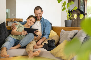 Mann umarmt Frau, die ein digitales Tablet hält, während er mit seinem Mops auf dem Sofa zu Hause sitzt - SBOF03233