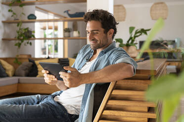 Lächelnder mittelgroßer erwachsener Mann, der auf ein digitales Tablet schaut, während er zu Hause auf der Couch sitzt - SBOF03234