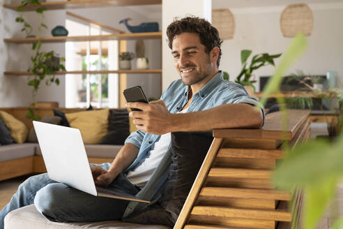 Smiling male freelance worker with laptop using smart phone while sitting couch in living room - SBOF03240