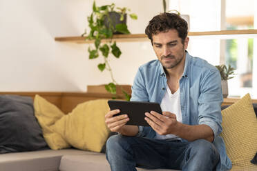 Ernster Mann, der ein digitales Tablet benutzt, während er zu Hause auf der Couch sitzt - SBOF03242