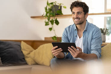 Lächelnder gut aussehender Mann, der ein digitales Tablet hält, während er zu Hause auf der Couch sitzt - SBOF03243