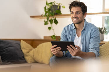 Smiling handsome man holding digital tablet while sitting on couch at home - SBOF03243