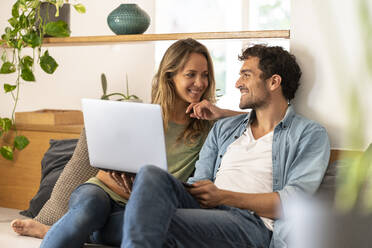 Smiling man with laptop looking at girlfriend while sitting on couch at home - SBOF03250