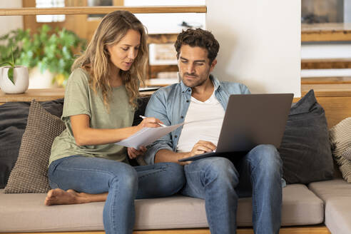 Girlfriend signing papers by man with laptop sitting on couch at home - SBOF03253