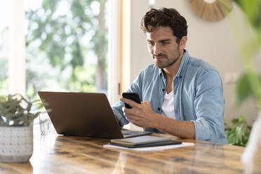 Serious male freelance worker holding mobile phone while using laptop at table - SBOF03256