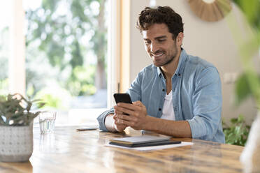 Lächelnder männlicher Freiberufler, der ein Mobiltelefon benutzt, während er am Tisch im Heimbüro sitzt - SBOF03257