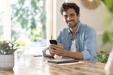 Smiling male freelance worker holding smart phone while sitting at table in home office - SBOF03258