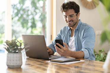 Lächelnder männlicher Freiberufler mit Brille, der ein Smartphone benutzt, während er mit einem Laptop im Büro zu Hause sitzt - SBOF03259