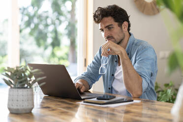 Concentrated male freelancer holding eyeglasses while working on laptop at table - SBOF03263