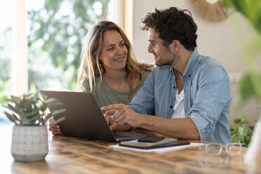 Smiling couple looking at each other while sitting in front of laptop at table - SBOF03264