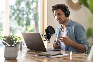 Männlicher Freiberufler, der bei der Aufnahme eines Podcasts zu Hause in ein Mikrofon spricht - SBOF03268