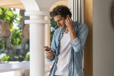 Mann, der ein Smartphone benutzt, während er über Kopfhörer am Türrahmen Musik hört - SBOF03276
