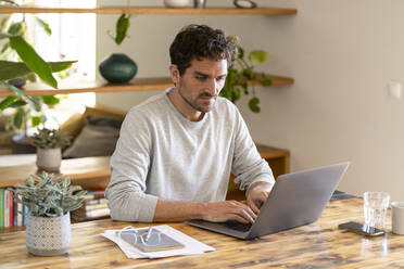 Concentrated male freelancer sitting at table working from home on laptop - SBOF03280