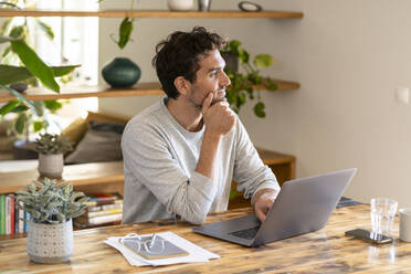 Contemplating freelance worker looking away while sitting with laptop at home office - SBOF03281