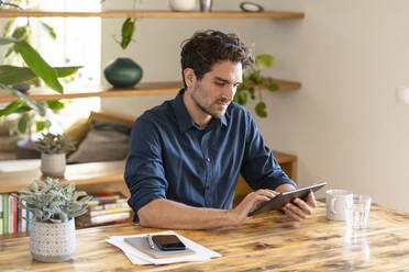 Pensive male freelancer using digital tablet while sitting at table in home office - SBOF03283