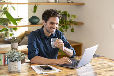 Lächelnder männlicher Freiberufler, der ein Glas Wasser in der Hand hält, während er im Heimbüro am Laptop arbeitet - SBOF03288