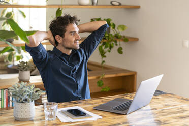 Relaxed male freelancer with hands behind head looking away while sitting in front of laptop at table - SBOF03290