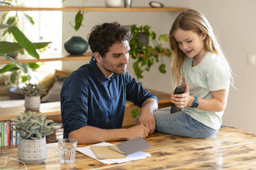 Girl showing mobile phone to father while sitting on table at home - SBOF03293