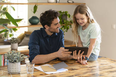 Happy father with digital tablet looking at daughter while discussing at home - SBOF03295