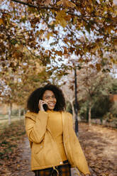 Happy Afro woman talking on smart phone in public park during autumn - TCEF01689