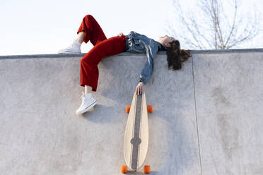 Niedriger Winkel Seitenansicht von müden weiblichen in trendigen Outfit mit Longboard in Beton Skatepark auf sonnigen Tag liegen - ADSF21930