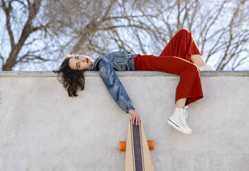 Niedriger Winkel Seitenansicht der müde Frau in trendigen Outfit mit Longboard in Beton Skatepark auf sonnigen Tag Blick auf die Kamera liegen - ADSF21932