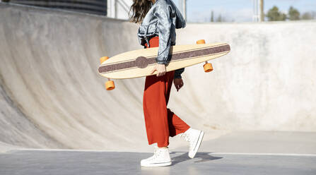 Seitenansicht einer abgeschnittenen, nicht erkennbaren, glücklichen jungen Frau in modischer Kleidung, die mit einem Longboard auf einem Skatepark aus Beton spazieren geht - ADSF21936
