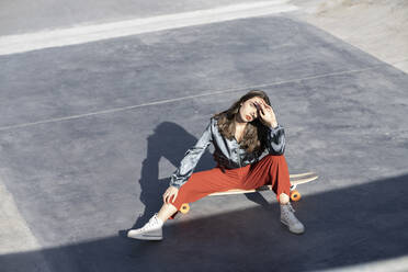 From above of young woman in stylish clothes touching forehead while sitting on longboard and looking at camera - ADSF21937