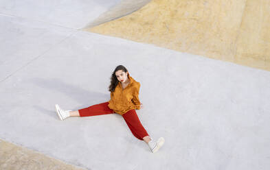 Von oben Ganzkörper junge Frau in stilvollem Outfit sitzt in Beton Skatepark und schaut in die Kamera im Sonnenlicht - ADSF21941