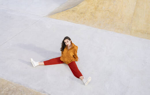 Von oben Ganzkörper junge Frau in stilvollem Outfit sitzt in Beton Skatepark und schaut in die Kamera im Sonnenlicht - ADSF21941