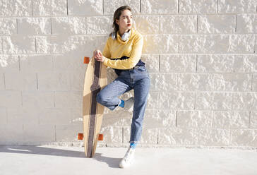 Young trendy female skateboarder with longboard standing with raised leg near rough wall while looking away - ADSF21943