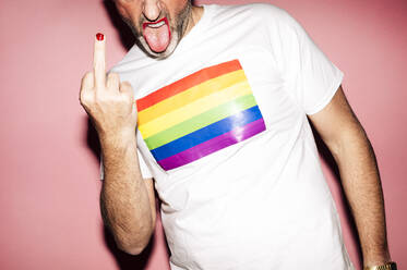 Crop unrecognizable unshaven gay with red lips in with t shirt with LGBT flag showing fuck gesture with tongue out against pink background - ADSF21950