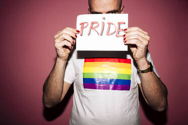 Rebellious bearded homosexual man with red lips and manicure making grimace while showing and covering face with paper with Pride text against pink background - ADSF21952