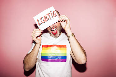 Crop rebellious bearded homosexual man with red lips and manicure making grimace while showing and covering face with paper with LGBTIQ text against pink background - ADSF21953