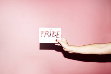 Crop unrecognizable homosexual person with manicure demonstrating white paper with PRIDE inscription against pink background - ADSF21955