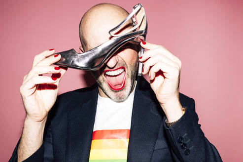 Eccentric bald bearded homosexual male with red lips and manicure in shirt with LGBT flag yelling and covering eyes with high heeled shoe against pink background - ADSF21957