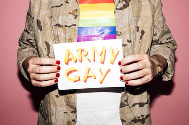 Crop anonymous bearded eccentric middle aged man in shirt with LGBT flag with manicure showing paper with Army Gay inscription against pink background - ADSF21960