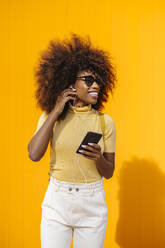 Zufriedene junge ethnische Frau mit Sonnenbrille und Afrofrisur, die im Internet auf ihrem Handy surft und Musik auf gelbem Hintergrund hört - ADSF21990