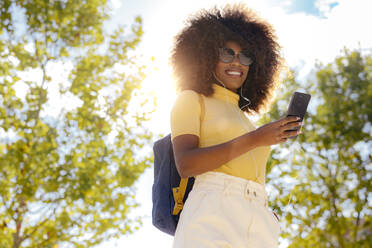 From below of glad ethnic female in earphones and sunglasses taking selfie on cellphone under blue sky in sunlight - ADSF21993