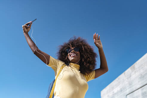 Von unten der fröhlichen ethnischen weiblichen in Kopfhörer und Sonnenbrille unter Selfie auf Handy unter blauem Himmel im Sonnenlicht - ADSF21995