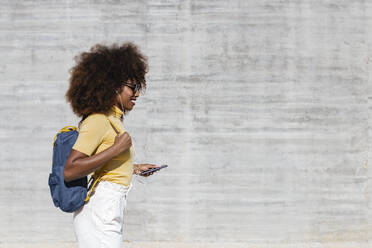 Glad ethnic female with Afro hairstyle and headphones walking near grey wall - ADSF21996