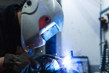 Seitenansicht eines nicht erkennbaren Mechanikers mit Schutzhelm, der einen Schweißbrenner beim Schweißen von Metallteilen in einer Werkstatt benutzt - ADSF22029