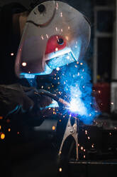 Seitenansicht eines nicht erkennbaren Mechanikers mit Schutzhelm, der einen Schweißbrenner beim Schweißen von Metallteilen in einer Werkstatt benutzt - ADSF22034