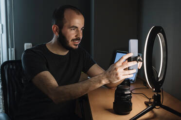 Male influencer adjusting mobile phone attached to illuminated circular strobe at studio - XLGF01345