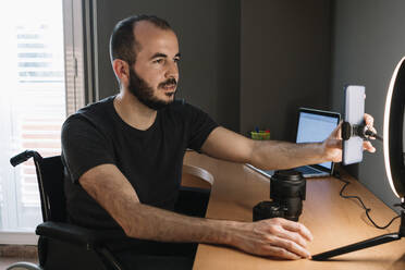 Disabled male influencer adjusting mobile phone attached with illuminated camera flash at studio - XLGF01349