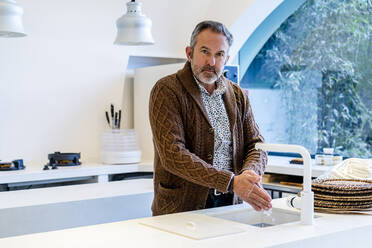 Mature man in sweater washing hands standing at kitchen sink at home - DLTSF01663