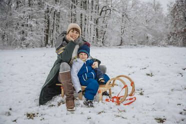 Mother and son sitting on sled at snowy field during winter - MFF07632