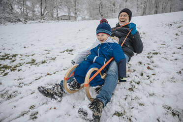 Fröhlicher Vater und Sohn beim Schlittenfahren auf einem verschneiten Hügel im Winter - MFF07633